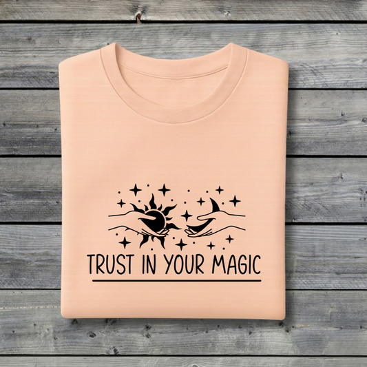 Folded peach sweatshirt with 'Trust in Your Magic' text and graphic on a wooden surface